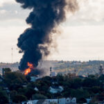 Large fire at an oil refinery seen from a distance. Only parts of the refinery visible behind the trees. Thick black smoke rises from the flames.