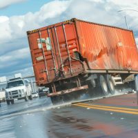 An intense photo of a container truck jackknifed across multiple lanes of traffic, blocking the roadway and causing chaos among other vehicles.